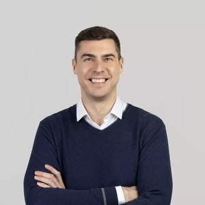 Vyras trumpais tamsiais plaukais dėvi baltus marškinius ir tamsiai mėlyną megztinį. Jis stovi sukryžiavęs rankas ir plačiai šypsosi žiūrėdamas į kamerą šviesiame fone.