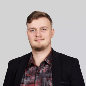 Portretinė nuotrauka – jaunas vyras trumpais šviesiais plaukais ir barzda, dėvintis juodą švarką ir margintus marškinius raudonai pilkais raštais. Jis žiūri į kamerą su lengva šypsena, fonas neutralus, šviesiai pilkas.