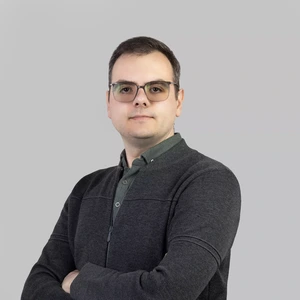 Portretinė nuotrauka – jaunas vyras su trumpais plaukais ir akiniais, vilkintis tamsų megztinį ir žalsvus marškinius, sukryžiavęs rankas žvelgia į kamerą šviesiame fone.