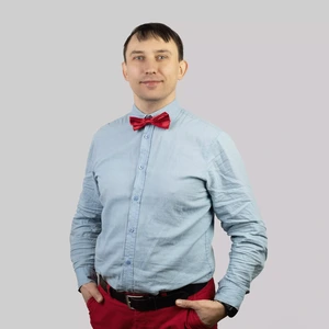 Portretinė nuotrauka – vyras trumpais tamsiais plaukais, vilkintis šviesiai mėlynus marškinius, ryšiantis ryškiai raudoną peteliškę ir dėvintis raudonas kelnes. Jo rankos įkištos į kišenes, jis žiūri tiesiai į kamerą, fonas neutralus, šviesiai pilkas.