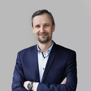 Portretinė nuotrauka – vyras su pilna barzda, vilkintis tamsų kostiumą ir šviesius marškinius su langeliais. Jis žiūri į kamerą su plačia šypsena, rankos sukryžiuotos, fonas neutralus, pilkšvas.