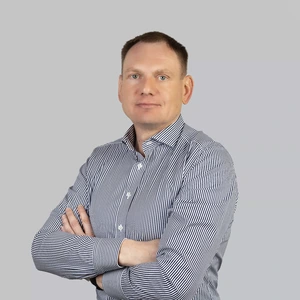 Portretinė nuotrauka – vyras trumpais tamsiais plaukais, dėvintis baltus marškinius smulkiomis mėlynomis juostelėmis. Jis stovi sukryžiavęs rankas ir žiūri į kamerą rimtu, pasitikinčiu žvilgsniu. Fonas neutralus, šviesiai pilkas.