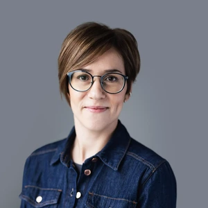 Portretinė nuotrauka – moteris trumpais rudos spalvos plaukais, su akiniais apvaliais juodais rėmeliais, vilkinti džinsinį marškinėlių tipo švarkelį. Ji švelniai šypsosi, fonas tamsiai pilkas, neutralus.