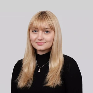 Portretinė nuotrauka – jauna moteris šviesiais tiesiais plaukais ir kirpčiukais, vilkinti juodą megztinį aukštu kaklu. Ji švelniai šypsosi, dėvi pakabuką, fonas neutralus, pilkšvas.