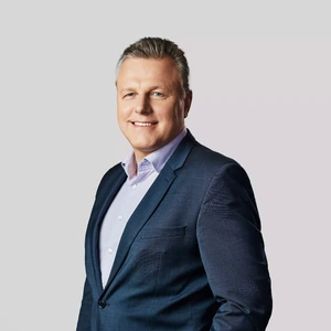Vyresnio amžiaus vyras trumpais šviesiais plaukais dėvi tamsų kostiumą ir šviesiai violetinius marškinius, šypsosi ir žiūri į kamerą šviesiame fone.
