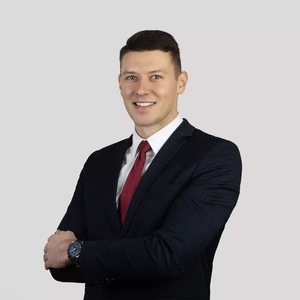 Portretinė nuotrauka – jaunas vyras trumpais plaukais, vilkintis tamsų kostiumą, baltus marškinius ir raudoną kaklaraištį, sukryžiavęs rankas šypsosi žvelgdamas į kamerą šviesiame fone. Ant riešo dėvi laikrodį.