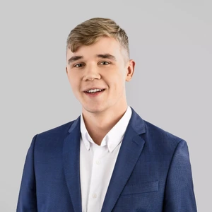 Jaunas vyras trumpais šviesiais plaukais dėvi mėlyną kostiumą ir baltus marškinius, šypsosi ir žiūri į kamerą šviesiame fone.