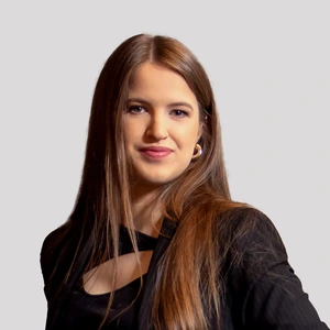 Ugnė Dirdaitė pozuoja portretinei nuotraukai šviesiame fone, dėvėdama juodą švarką ir subtilius auskarus.