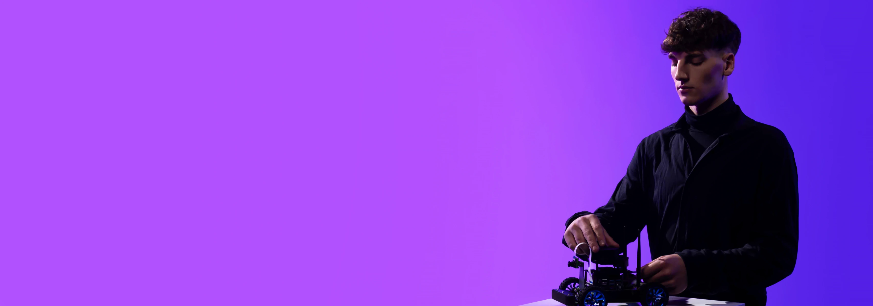 Jaunuolis violetiniame fone valdo mažą mobilų robotą, tinkamą studijoms apjungiančioms technologijas ir verslą.