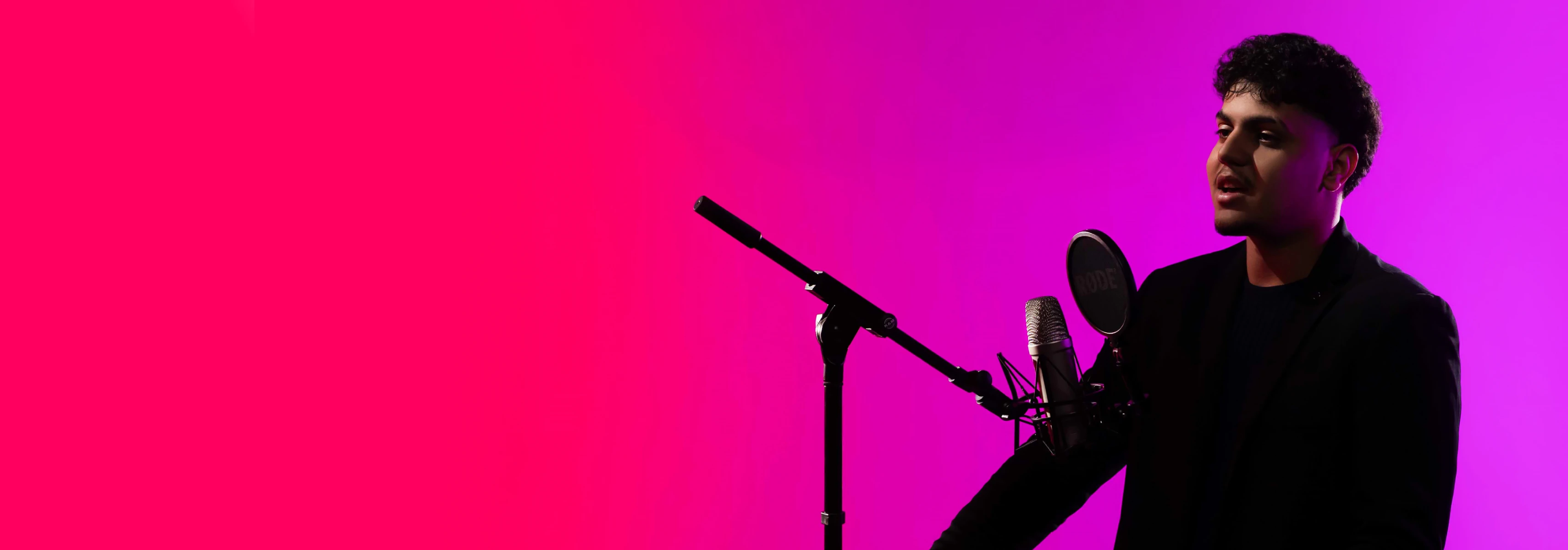 Vaikinas violetiniame fone kalba į mikrofoną su stoveliu, tinkamą socialinių, humanitarinių mokslų ar menų krypties veikloms.