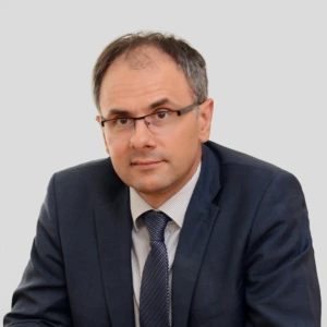 Portretinė nuotrauka – vidutinio amžiaus vyras trumpais, šiek tiek žilstelėjusiais plaukais ir akiniais. Jis dėvi tamsiai mėlyną kostiumą, baltus dryžuotus marškinius ir tamsų, subtiliai raštuotą kaklaraištį. Žiūri tiesiai į kamerą neutraliu žvilgsniu. Fonas vientisas, šviesiai pilkas.