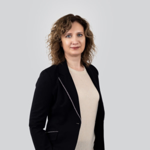 Portretinė nuotrauka – vidutinio amžiaus moteris banguotais, pečius siekiančiais šviesiai rudais plaukais. Ji dėvi šviesų megztinį ir juodą švarką su subtiliomis siūlėmis. Žiūri tiesiai į kamerą ramiu, pasitikinčiu žvilgsniu. Fonas neutralus, šviesiai pilkas.