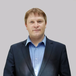 Portretinė nuotrauka – šviesiaplaukis vyras vidutinio amžiaus, trumpais plaukais. Jis dėvi šviesiai mėlynus marškinius ir tamsiai mėlyną kostiumą. Žiūri tiesiai į kamerą ramiu veidu. Fonas neutralus, šviesiai pilkas.