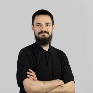 Jaunas vyras trumpais tamsiais plaukais, su ūsais ir barzda, vilkintis tamsiais polo marškinėliais, rankos sukryžiuotos priekyje, profesionaliai besišypsantis, fotografuotas neutraliame fone.