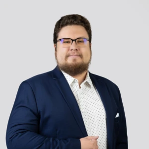 Jaunas vyras trumpais tamsiais plaukais, su akiniais ir barzda, vilkintis tamsiai mėlyną švarką ir šviesius raštuotus marškinius, ramios ir profesionalios išraiškos, fotografuotas neutraliame fone.