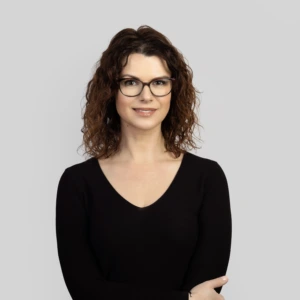 Jauna moteris rudais, banguotais, pečius siekiančiais plaukais, su tamsiais akiniais, vilkinti juodą suknelę, rankos sukryžiuotos priekyje, profesionaliai besišypsanti, fotografuota neutraliame fone.