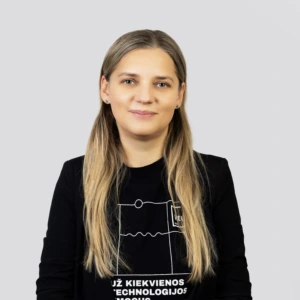 Eglė Januškienė Jauna šviesiaplaukė moteris su tiesiais, ilgais plaukais, vilkinti juodą megztinį, profesionaliai besišypsanti neutraliame fone.