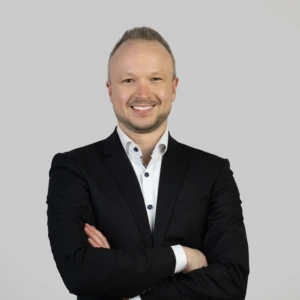 Jaunas vyras trumpais tamsiais plaukais, vilkintis tamsų prasegtą švarką ir šviesius marškinius, rankos sukryžiuotos priekyje, profesionaliai besišypsantis, fotografuotas neutraliame fone.