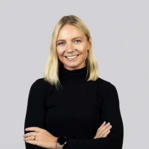 Jauna šviesiaplaukė moteris su tiesiais, pečius siekiančiais plaukais, vilkinti juodą megztinį aukštu kaklu, rankas sukryžiavusi, profesionaliai besišypsanti neutraliame fone.