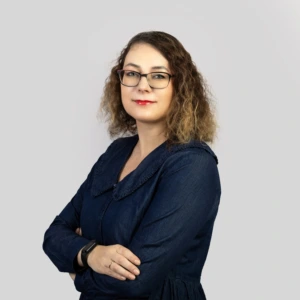 Jauna moteris rudais, pečius siekiančiais plaukais, vilkinti tamsiai mėlyną suknelę, rankos sukryžiuotos priekyje, profesionaliai besišypsanti, fotografuota neutraliame fone.