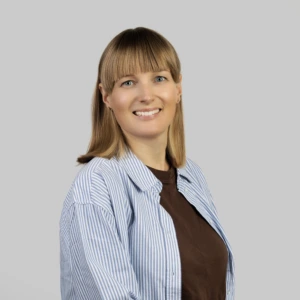 Jauna šviesiaplaukė moteris su tiesiais, pečius siekiančiais plaukais, vilkinti mėlynus, dryžuotus, prasegtus marškinius, rudą palaidinę, profesionaliai besišypsanti neutraliame fone.