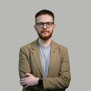 Jaunas vyras trumpais tamsiais plaukais, trumpa barzda su akiniais tamsiais rėmeliais, vilkintis šviesiai rudą švarką ir pilkus marškinėlius, rankos sukryžiuotos priekyje, profesionalios išraiškos, fotografuotas neutraliame fone.