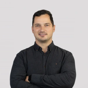 Jaunas vyras trumpais tamsiais plaukais, vilkintis tamsiais marškiniais su prasegta viršutine saga, rankos sukryžiuotos priekyje, profesionalios išraiškos, fotografuotas neutraliame fone.