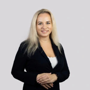 Jauna šviesiaplaukė moteris su tiesiais, pečius siekiančiais plaukais, vilkinti juodą švarką ir šviesią palaidinę, rankas sukryžiavusi, profesionaliai besišypsanti neutraliame fone.