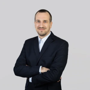 Jaunas vyras trumpais tamsiais plaukais, su ūsais ir barzda, vilkintis tamsų švarką ir šviesius marškinius, rankos sukryžiuotos priekyje, profesionaliai besišypsantis, fotografuotas neutraliame fone.