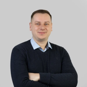 Jaunas vyras trumpais tamsiais plaukais, vilkintis tamsų megztinį ir šviesius marškinius, rankos sukryžiuotos priekyje, profesionaliai besišypsantis, fotografuotas neutraliame fone.