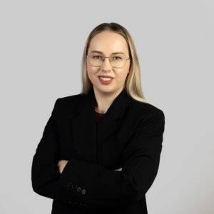 Jauna šviesiaplaukė moteris su tiesiais, pečius siekiančiais plaukais, vilkinti juodą švarką, rankas sukryžiavusi, profesionaliai besišypsanti neutraliame fone.