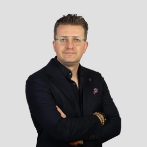 Jaunas vyras trumpais tamsiais plaukais, su akiniais, vilkintis tamsų švarką ir tamsius marškinius, rankos sukryžiuotos priekyje, profesionali veido išraiška, fotografuotas neutraliame fone.