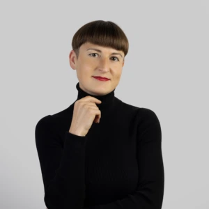 Jauna moteris rudais, trumpais plaukais, vilkinti juodą palaidinę aukštu kaklu, vieną ranką pakėlusi prie smakro, kitą nuleidusi, profesionaliai besišypsanti neutraliame fone.