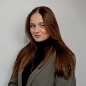 Portretinė nuotrauka – Roberta Kybartaitė. Jauna moteris ilgais tiesiais rudais plaukais. Ji dėvi juodą aukštu kaklu megztinį ir pilkai žalsvą švarką. Moteris šiek tiek pasisukusi į šoną, tačiau žvilgsnis nukreiptas į kamerą, veide matoma lengva šypsena. Fonas šviesus ir neutralus, studijinio tipo.