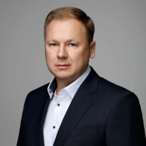 Edmundas Kivilša Edmundo Kivilšos portretas pilkame fone. Jis vilki tamsų kostiumo švarką ir baltus marškinius bei žvelgia tiesiai į kamerą.