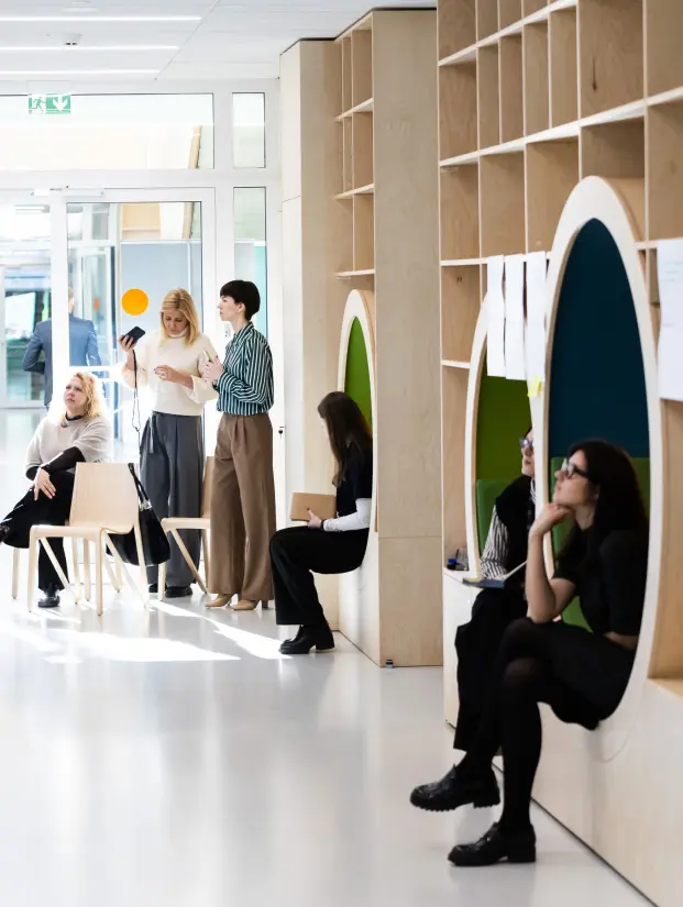 Žmonės bendrauja ir dirba šviesioje modernioje studijų erdvėje su medinėmis lentynomis ir integruotomis sėdėjimo nišomis. Neformali aplinka iliustruoja bendradarbiavimu ir idėjų mainais grįstą KTU MBA studijų patirtį.