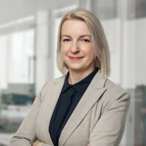 Ramunė Šličienė Ramunės Šličienės portretas šviesioje modernioje darbo aplinkoje. Ji vilki šviesiai pilką švarką ir tamsius marškinius. Sukryžiavusi rankas šypsosi ir žvelgia į kamerą.