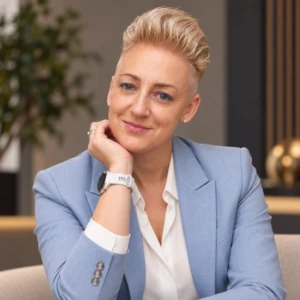Živilė Čepkauskaitė-Bagdonienė Živilės Čepkauskaitės-Bagdonienės portretas modernioje interjero aplinkoje. Ji vilki šviesiai mėlyną švarką ir baltus marškinius. Galvą parėmusi ranka šypsosi ir žvelgia į kamerą.
